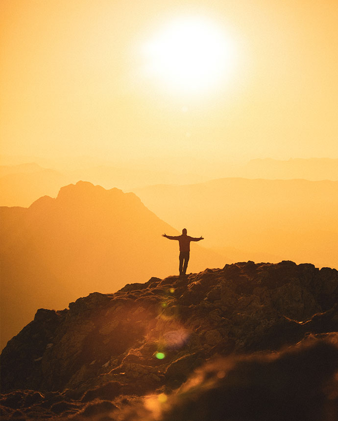 Eine Person steht mit ausgestreckten Armen auf einem Gipfel, während die Sonne in warmen Gelbtönen über die Berge strahlt. Das Bild symbolisiert Freiheit, Sicherheit und Zuversicht und spiegelt die Leistungen der Voigt Versicherungen wider, die durch ihr Beamtencare-Servicepaket für finanzielle und persönliche Absicherung sorgen.