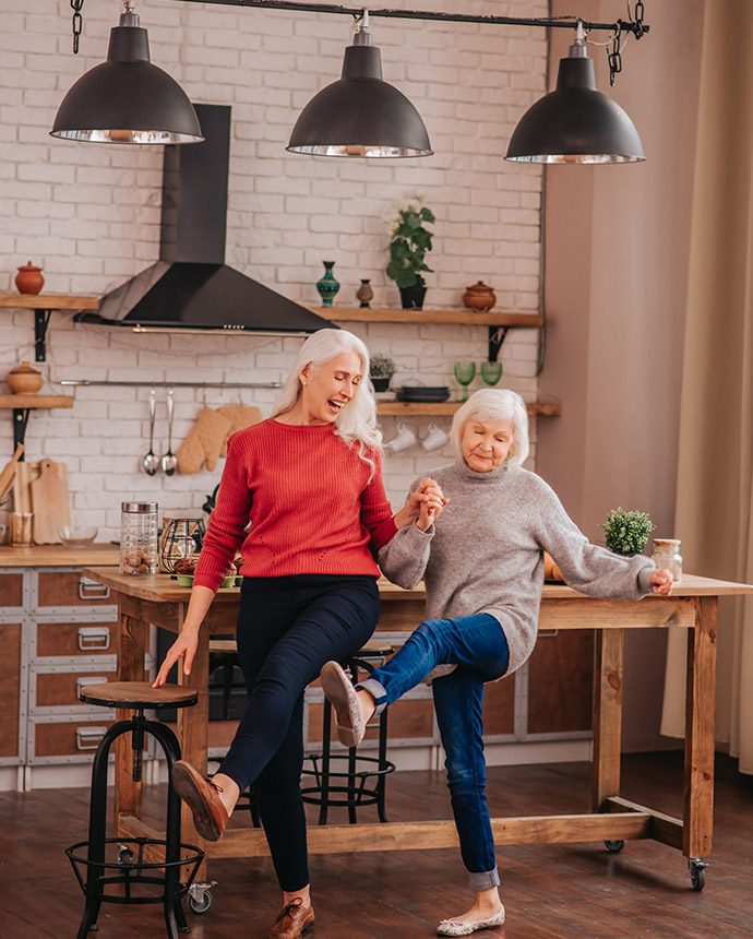 Zwei Frauen, eine jüngere und eine ältere, tanzen gemeinsam in einer gemütlichen, modern eingerichteten Küche. Dieses Bild symbolisiert Lebensfreude und familiären Zusammenhalt, den Voigt Versicherung durch eine zuverlässige Wohngebäudeversicherung absichert. Genießen Sie unbeschwerte Momente – mit unserer Unterstützung!