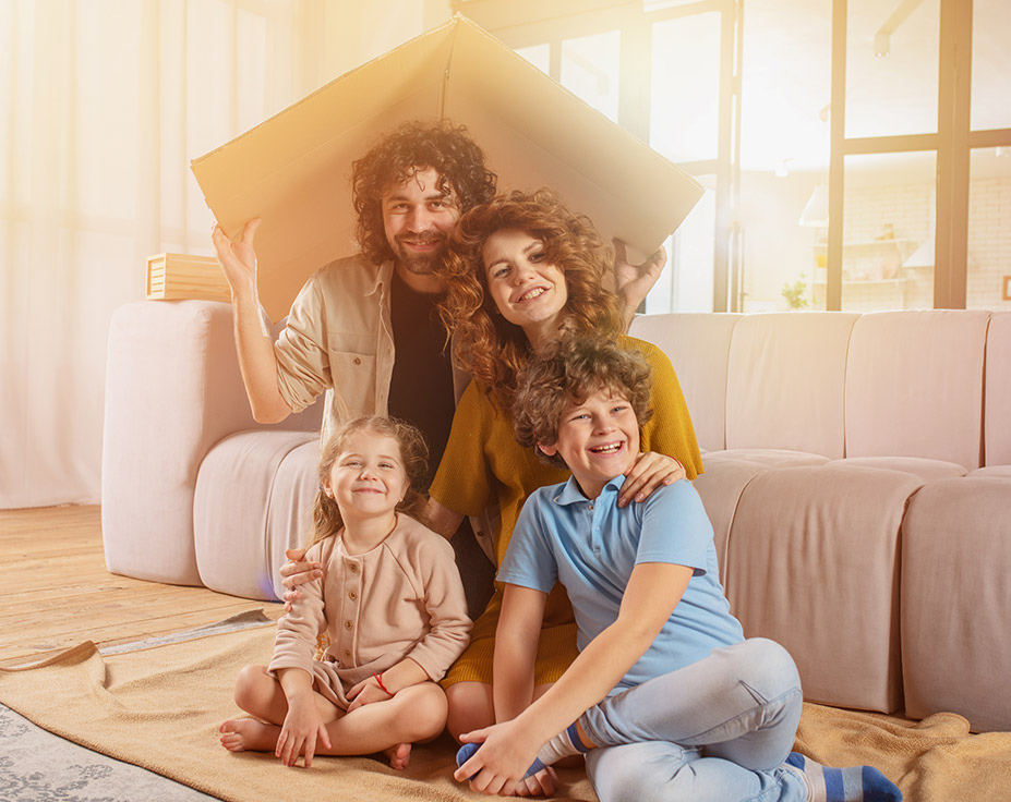 Eine glückliche Familie sitzt in einem gemütlichen Wohnzimmer. Die Eltern halten gemeinsam einen Pappkarton über ihre Köpfe, als symbolischer Schutz. Im Vordergrund lachen die beiden Kinder, ein Junge im blauen T-Shirt und ein Mädchen im rosa Pullover. Die Szene ist in warmes Licht getaucht und vermittelt ein Gefühl von Geborgenheit und Sicherheit – passend für die Themen Wohngebäude- und Hausratsversicherung bei Voigt Versicherung.